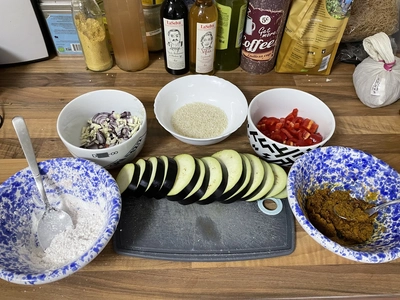 Arbeitsplatte mit Schüsseln, in denen Paprikastreifen, gehackter Knoblauch und Zwiebeln, Pesto und Reis liegen. Inmitten dessen ist ein Brettchen mit Auberginen